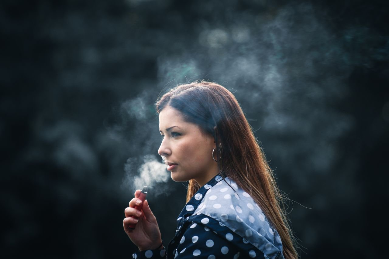 A woman with brown hair using an e-cigarette, surrounded by smoke in an outdoor setting.