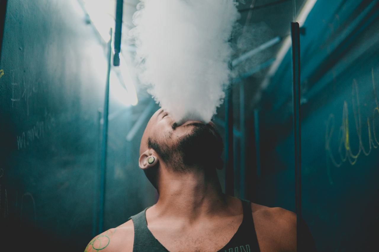 A bearded man in a tank top exhales smoke in a moody indoor environment.